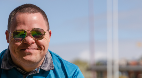 An NDIS participant is smiling at the camera. He's wearing a blue shirt, sunglasses and is sitting outside. 