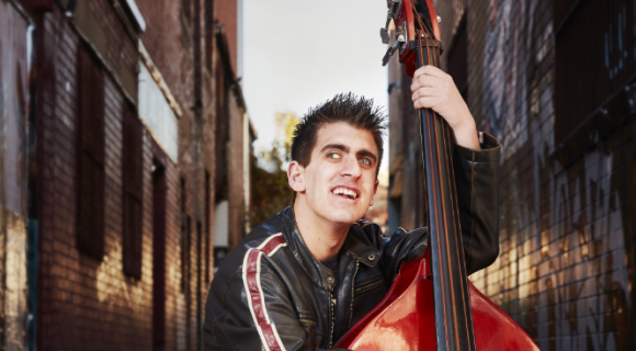 Ricky is standing in a laneway playing his cello. He is wearing a black leather jacket and his hair is spiked.