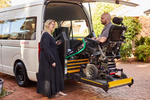James is in his electric wheelchair and is being lifted into the back of a van by an eletric platform. His support worker is standing next to him using the controls. James is wearing a grey shirt and is looking at his support worker.