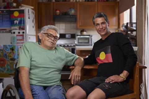 John and his support worker Eleisha are sitting at a dining table with a kitchen in the background. John is wearing a green t-shirt and jeans, and has one arm resting on the table. Eleisha is wearing a black shirt with the Aboriginal flag on the front. Both are looking at the camera and smiling.