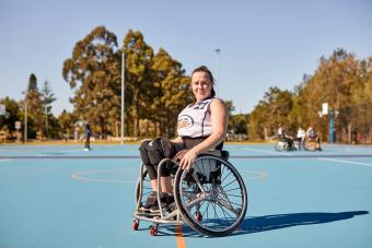 Hannah is sitting in her wheelchair in activewear. She is on a netball court and is looking at the camera. There are people and trees in the background.